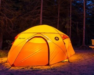 A brightly lit orange Marmot tent glowing at night in a dark forest campsite, suggesting cozy camping near Montreal.