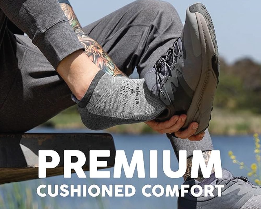 A person wearing a grey Balega Hidden Comfort sock and dark grey athletic shoes, sitting on a bench with a lake in the background, highlighting "PREMIUM CUSHIONED COMFORT."