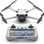 A white DJI Mini 3 Pro drone with its four propellers and an integrated camera facing forward, positioned above its gray remote controller. The remote controller features a built-in screen displaying a panoramic winter mountain landscape at sunset, with joysticks and control buttons visible. This setup represents a leading option for the Best Drone Camera.