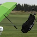 A Titleist golf ball on a tee, a black Titleist golf bag, and a green umbrella on a lush golf course with a treeline in the background.