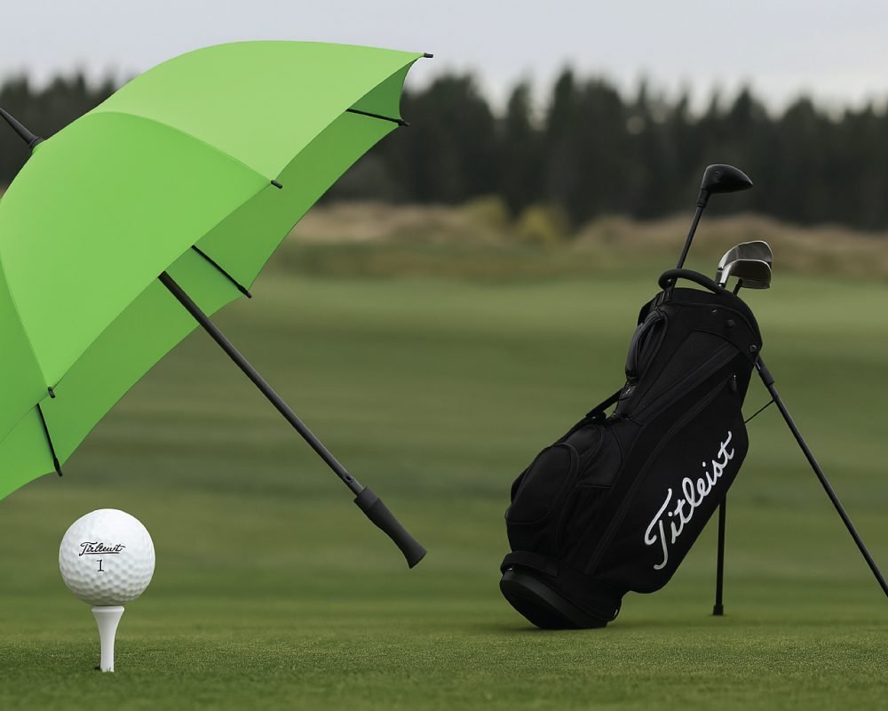 A Titleist golf ball on a tee, a black Titleist golf bag, and a green umbrella on a lush golf course with a treeline in the background.