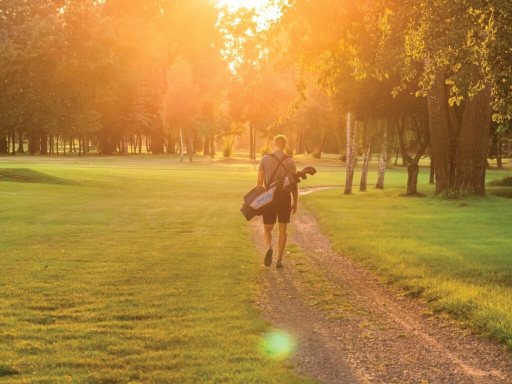A golfer walking down a path on a sunny day at a park golf course, carrying a golf bag over his shoulder.