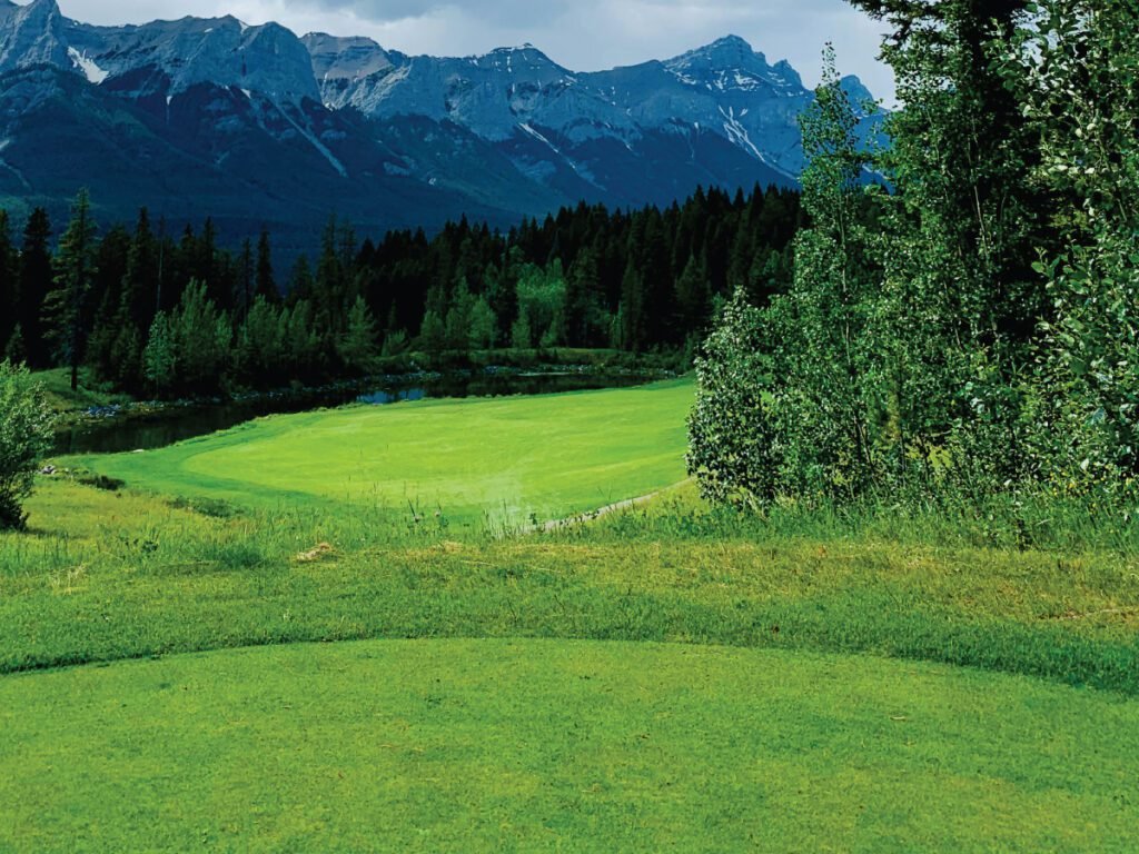 A pristine golf course fairway in Jasper National Park, Alberta, with a small pond, a line of dense forest, and rugged, snow-dusted mountains under a cloudy sky.