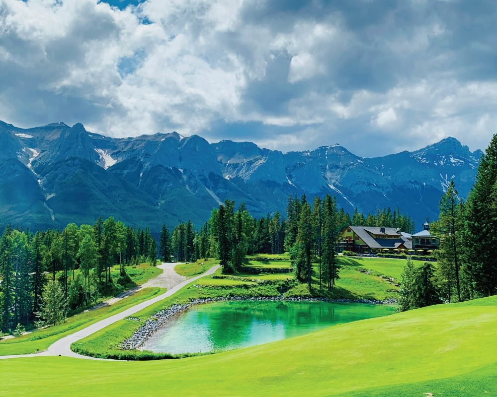  A scenic view of a beautiful provincial park golf course with a green pond, a lodge, and majestic, snow-capped mountains in the background, a true hidden gem for golfers.